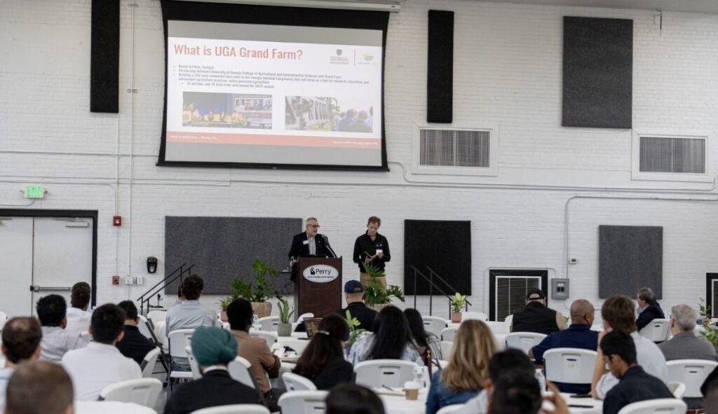 Two speakers stand at the front of a room addressing a group of seated conference participants.